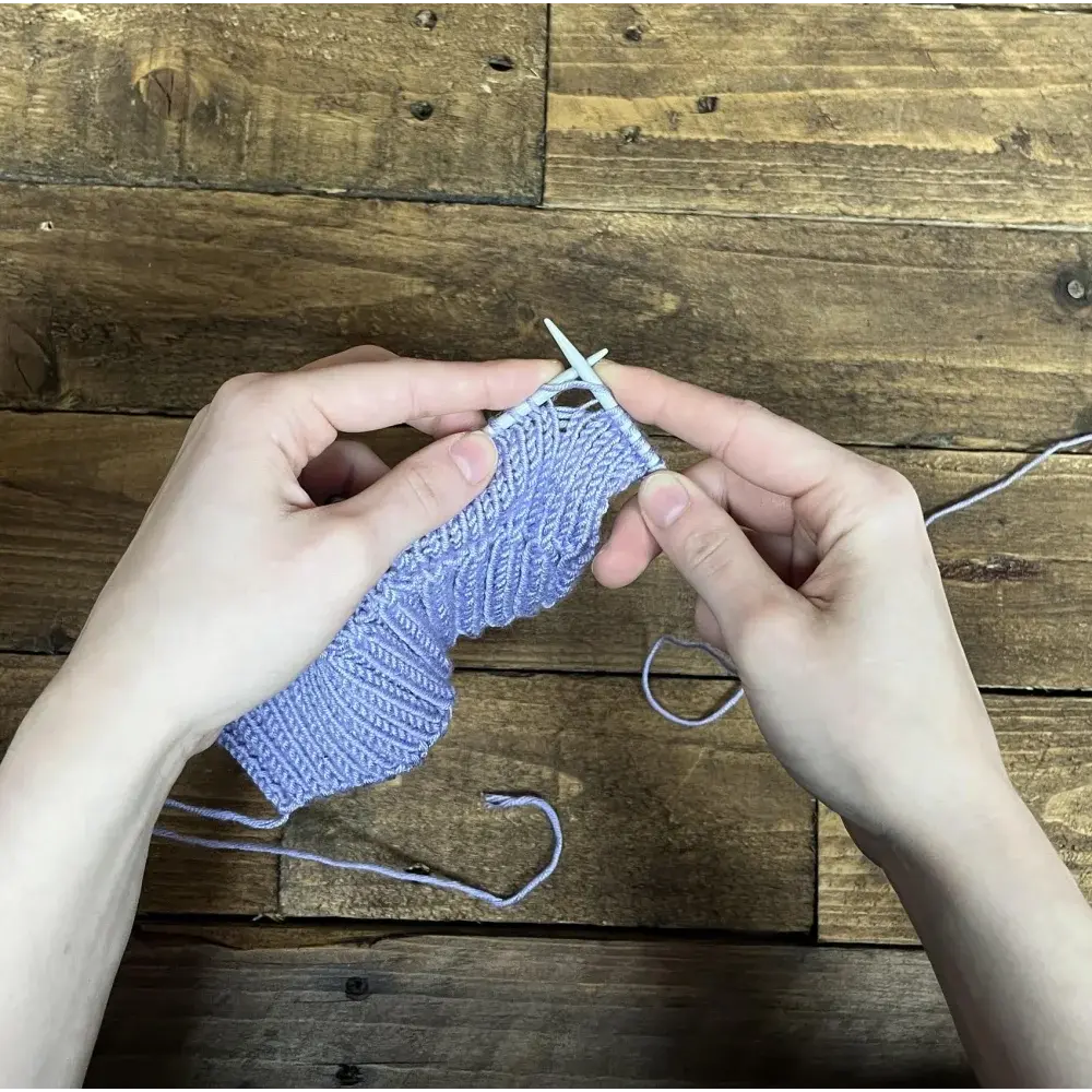 Hands knitting a light purple ribbed fabric.