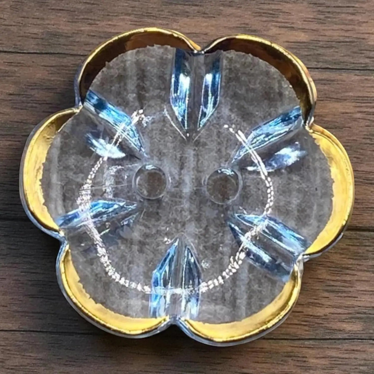 KnitForLess 13mm baby blue flower-shaped acrylic button with a gold rim and two-hole design on a wooden surface.