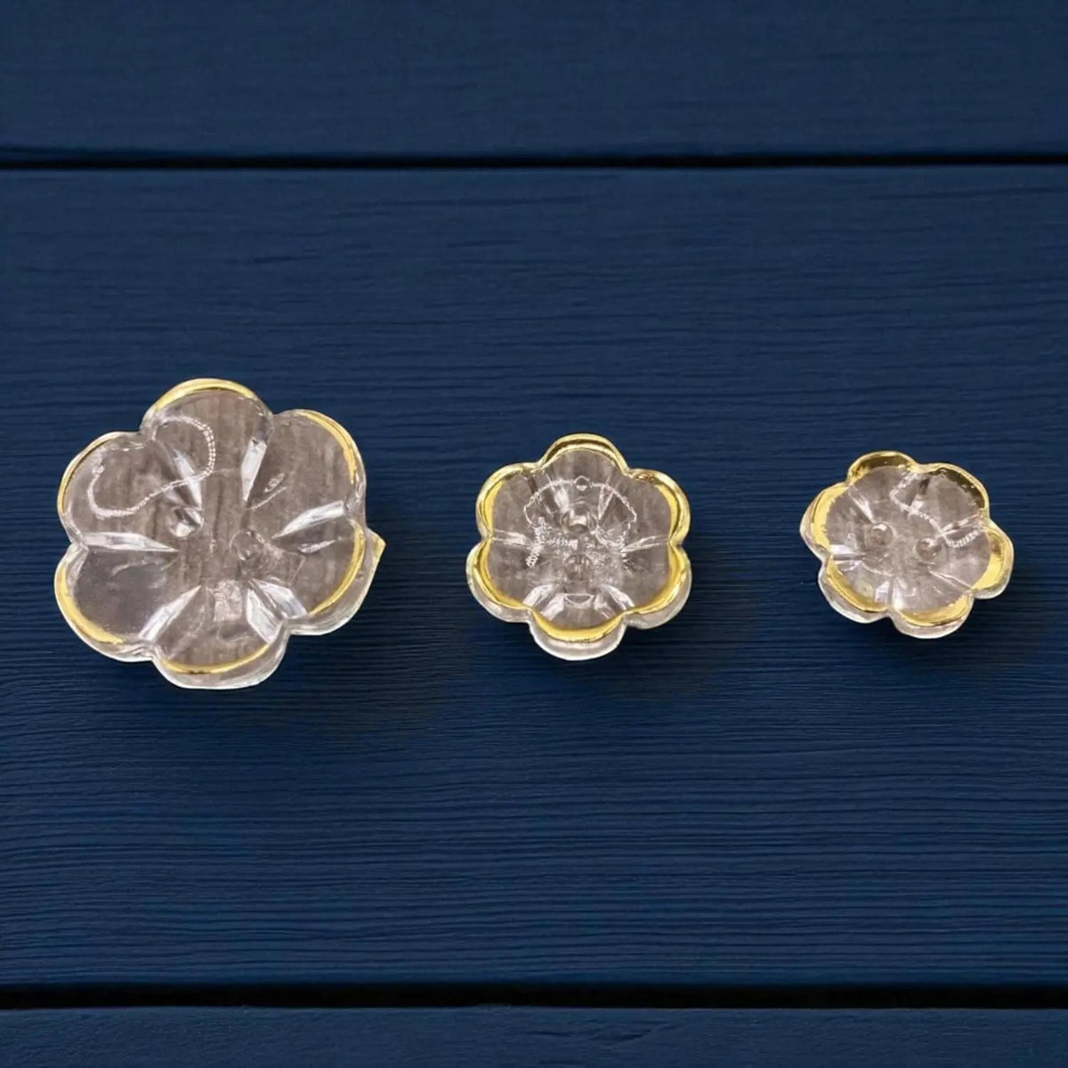 KnitForLess clear acrylic flower-shaped buttons with gold rims in various sizes on a dark blue wooden surface