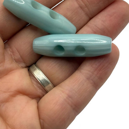 KnitForLess light blue barrel toggle buttons with a smooth glossy finish held in a hand for knitwear and coats.