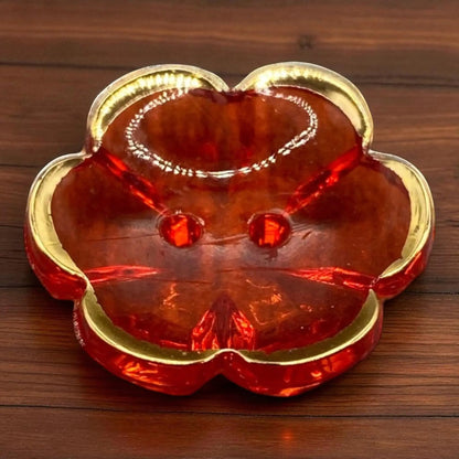 KnitForLess red flower-shaped acrylic button with a gold rim and two holes, resting on a dark wood surface.